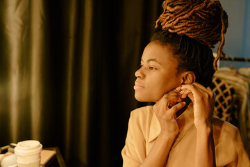 African American woman adjusting her earrings while looking away. Wearing beige top with dreadlocks styled in a high updo, she appears focused and tranquil