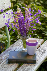 Morning cocoa with purple lupine flowers and book on rustic garden bench.