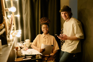 Smiling makeup artist sitting at her vanity table full with tools and products. A casual observer standing beside holding a cellphone and observing the scene