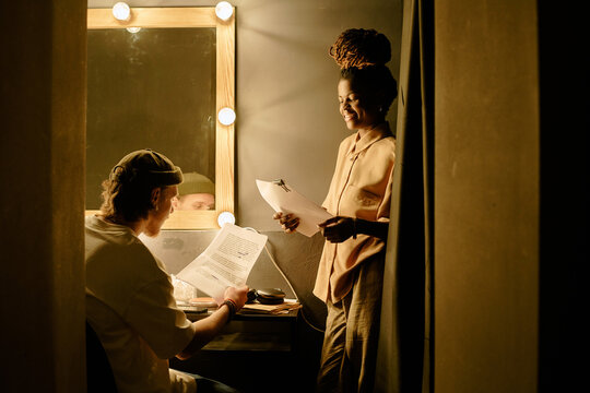 Two people in dressing room collaborating on theater script. One person seated at dressing table under warm lighting, while another person standing beside holding script pages