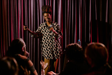 Person delivering stand-up comedy routine on stage holding microphone engaging with diverse audience at comedy club beaming bright smile energetic delivery of jokes