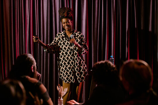 Person delivering stand-up comedy routine on stage holding microphone engaging with diverse audience at comedy club beaming bright smile energetic delivery of jokes