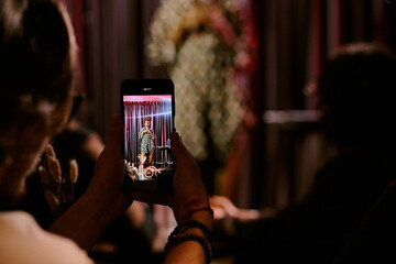 Audience member capturing comedian performing on stage using mobile phone at live event with focus on screen and blurred background, emphasizing entertainment atmosphere
