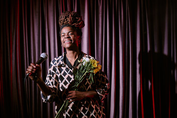 Obraz premium African American performer smiling while holding microphone and bouquet of flowers on stage during performance with long dreadlocks and patterned outfit