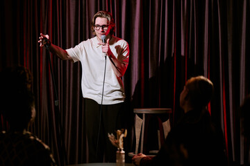 Comedian engaging audience with humor and wit during stand-up performance on stage. Microphone held...