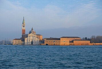 San Giorgio Maggiore island in Venice, with its historic Benedictine church and bell tower, bathed in warm sunlight. Architecture contrasts with deep blue waters of Venetian lagoon, picturesque scene.