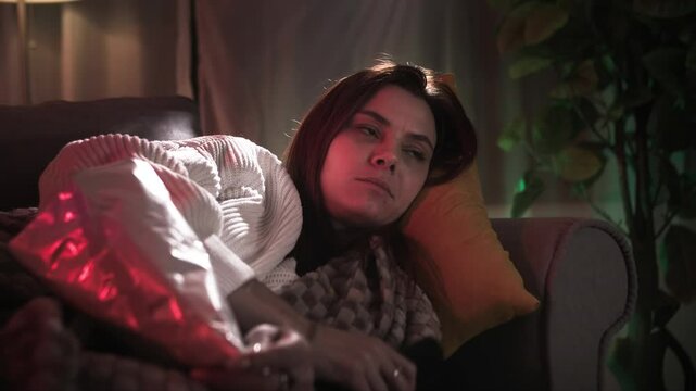 pretty redhead woman lying on pillow and using tv remote control, eating potato chips in warm backlight