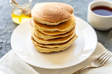 A pile of pancakes on a white plate with a napkin on a gray background with honey and a cup of tea.
