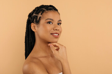 Side profile portrait of a young African American woman with braided hair and flawless skin, smiling softly while resting her hand under her chin. She is wrapped in a towel, posing against a beige