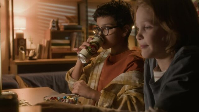 Side view shot of cute young boy wearing thick glasses and drinking soda while playing video games with friend using PC computer in 90s setting
