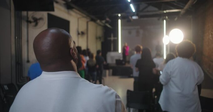 Wide shot of church service with congregation standing and worshipping, back view of people focused on stage, modern church interior, unity in faith