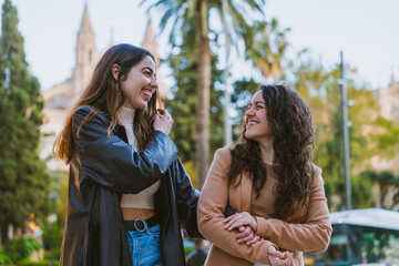 Fototapeta premium Two Cheerful Women Laughing Together in Park Setting. Smiling friends enjoy a fun and candid moment outdoors, surrounded by greenery and historic architecture.