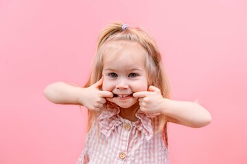 A little child girl laughs and fools around, stretching her mouth with her hands on a pink isolated...