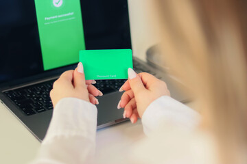 Hand holding a green discount card near a laptop. Online shopping, special offer, payment, and remote purchase.