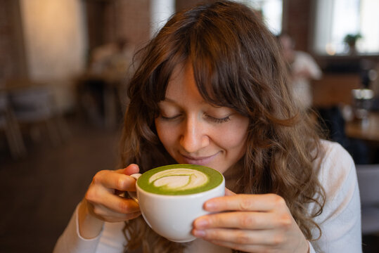 Closed eyes and a gentle smile reflecting pure joy, a young woman savoring the rich aroma of a matcha latte in a cozy, inviting cafe atmosphere, embracing moments of relaxation and tranquility