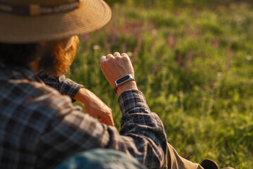 A man enjoys the outdoors, checking his smartwatch in a lush green setting illuminated by gentle sunlight.