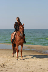 Pretty young brunette woman in black dress riding a horse at the sea