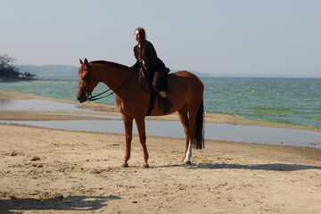 Pretty young brunette woman in black dress riding a horse at the sea