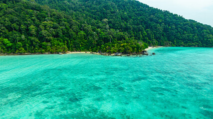 A breathtaking aerial view of a secluded Surin islands with crystal-clear turquoise waters, surrounded by lush green vegetation and corel reef. The pristine white sand and vibrant ocean hues