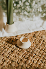A white candle holder sits on a rug next to a green candle holder. The candle holders are placed on a white cloth