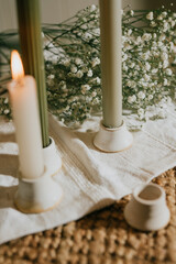 A white candle is lit next to a bunch of white flowers. The candle is surrounded by a white cloth and the flowers are placed on top of it. Concept of warmth and tranquility
