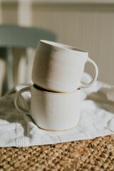 Two white coffee mugs are stacked on top of each other on a table. The table is covered with a white cloth and a brown rug. Concept of warmth and comfort, as the mugs