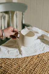 A hand holding a white mug on a table with a white cloth. The mug is placed on a rug