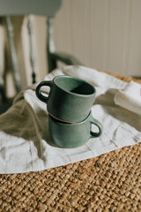 Two green coffee cups are sitting on a white cloth. The cups are placed on a rug, and the cloth is on a chair. The scene gives off a cozy and relaxed vibe