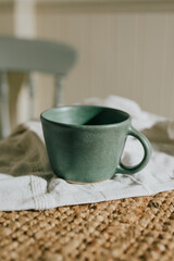 A green mug sits on a table next to a chair. The mug is made of ceramic and has a unique design. The table is covered with a white cloth, and the chair is positioned behind the mug