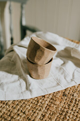 Two cups are sitting on a white cloth. The cups are made of clay and have a rustic appearance. The...