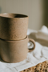 Two cups with a brown color and a rough texture. They are placed on a white cloth. The cups are not filled with anything