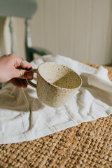A hand holding a white cup with a pattern on it. The cup is sitting on a table with a white cloth underneath it