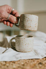 Two white ceramic cups with a design on them. The cups are placed on a white cloth. The cups are held by a person's hand