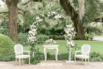 Obraz premium Elegant outdoor wedding ceremony setup. Lush greenery and white flowers create a romantic archway, small table, and chairs