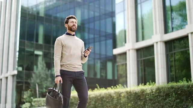 Confident handsome businessman walking outside modern glass building, holding phone and bag. Represents urban lifestyle, work-life balance, mobility, and technology in professional environment