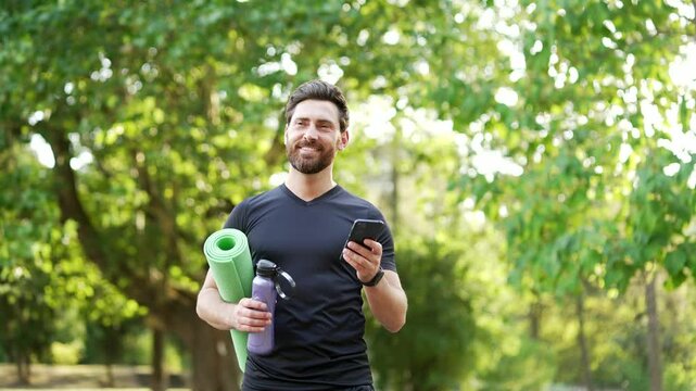 Sportsman engages in fitness by walking in park with yoga mat and water bottle, checking phone for workout updates. Smiling bearded athletic returning from training. Active lifestyle, healthy routine
