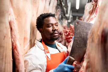 Confident black man butcher working in slaughterhouse cutting raw meat.