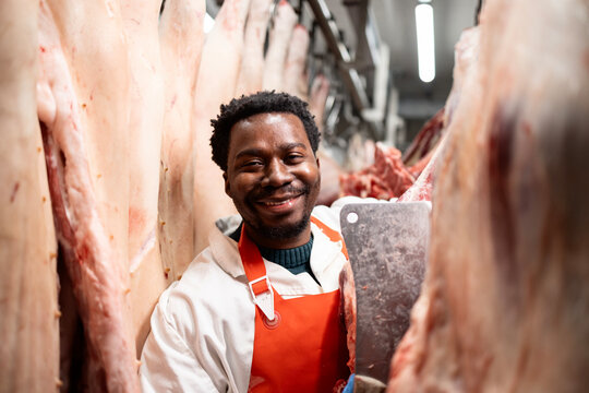 Portrait of smiling butcher working in slaughterhouse cutting hanging raw meat. - Powered by Adobe