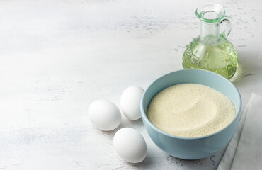 Baking ingredients: light blue bowl with semolina, fresh white eggs, a glass bowl of granulated sugar, and a bottle of vegetable oil on a white kitchen surface