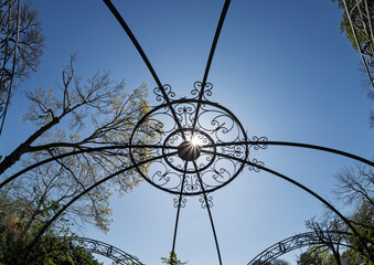 View looking up through a decorative iron gazebo roof toward a bright sun in a cloudless sky framed...