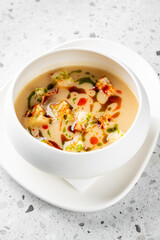 Creamy soup with croutons, garnished with green and red sauces, served in a white bowl on a white plate with a napkin, placed on a stone surface. Vibrant and appetizing presentation.