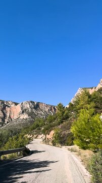 Remote, winding road curves toward the iconic limestone cliffs of Leonidio, Greece. Bright blue sky contrasts with the rugged orange rock that rises above rugged terrain and Mediterranean greenery.