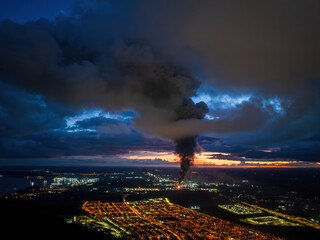 Aerial drone view of an industrial fire in Puerto Montt, Chile, with a thick smoke column rising over the city, dramatic clouds, and post-sunset light illuminating the urban landscape.