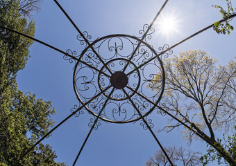 View looking up through a decorative iron gazebo roof toward a bright sun in a cloudless sky framed...