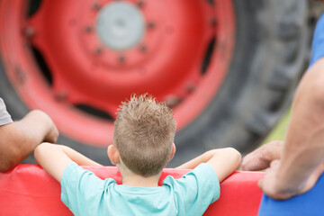 Young boy observes large tractor tire at outdoor agricultural event with adults nearby in a rural...
