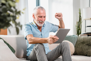 Overjoyed elderly Caucasian man relax at home look at tablet screen celebrate online lottery win....