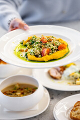 A hand holds a white plate with a grain salad, cherry tomatoes, herbs, and yellow hummus, surrounded by soup, bread, and other dishes on a wooden table. Fresh and colorful meal.