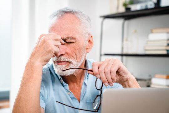 Fatigued mature old man taking off glasses suffering from tired dry irritated eyes after long computer use, senior middle aged male feels eye strain problem or blurry vision working on laptop at home