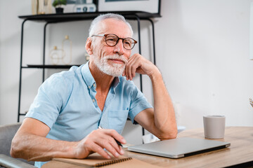 Fototapeta premium Portrait of happy handsome mature gray-haired caucasian freelancer or manager, stylishly dressed, sitting at the table, taking a break from work, dreaming about vacation, looking away, hands joining
