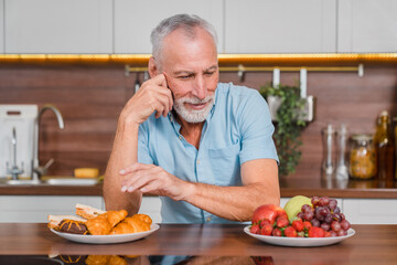 A senior man sitting in kitchen ponders a choice between a plate of fresh fruits and sugary pastries, suggesting themes of healthy eating and decision-making in diet. Nutrition diet concept
